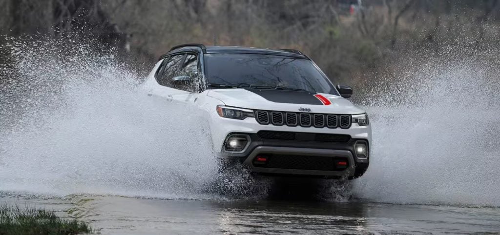 A white Jeep Compass with a black roof and red hood stripe drives through water, spraying it outwards dramatically.