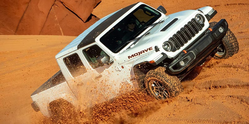 White Jeep Gladiator Mojave pickup truck driving up a steep red dirt path, kicking up debris.