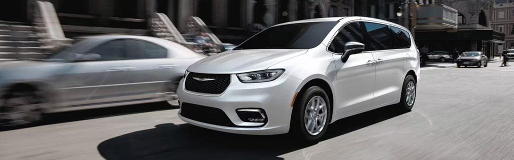 A white minivan drives down a city street with other cars and pedestrians blurred in the background.