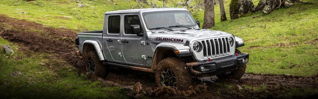 A silver Jeep Gladiator Rubicon pickup truck muddies a dirt trail through green grass and trees.