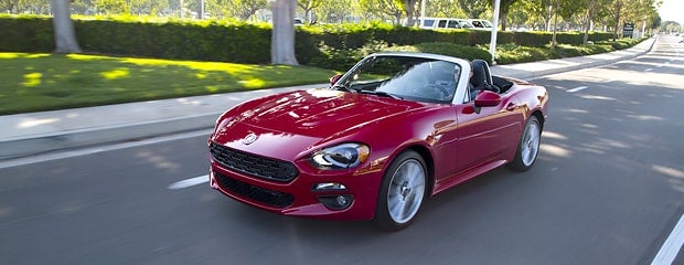 Red Fiat 124 Spider convertible, with the top down, driving on a road lined with green bushes and trees.