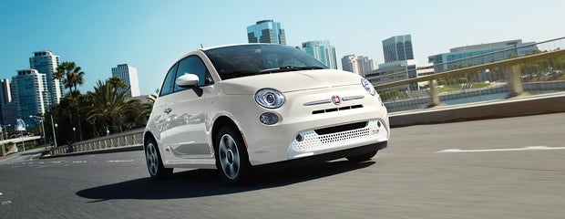 White Fiat 500e driving on a bridge, with city buildings and palm trees in the background.