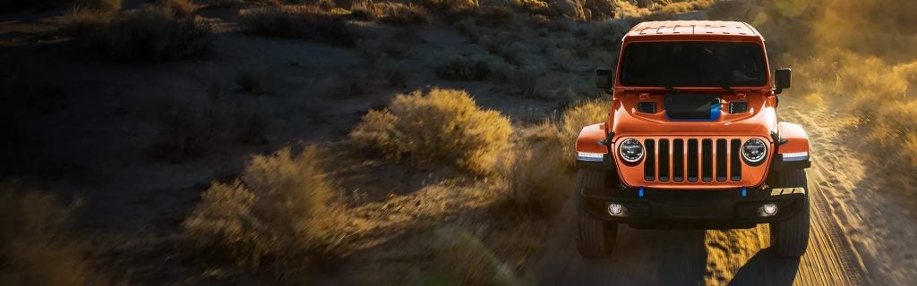An orange Jeep Wrangler 4xE offroading on a dirt path, golden sunlight illuminating the dust and arid landscape.