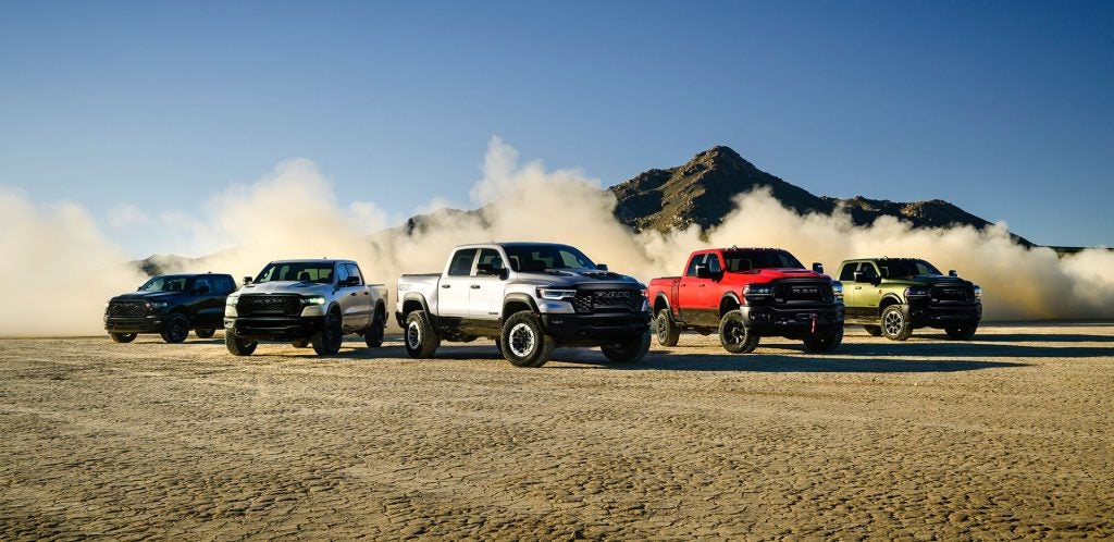 Five Ram TRX trucks, in different colors, kicking up dust in a desert landscape under a clear blue sky.