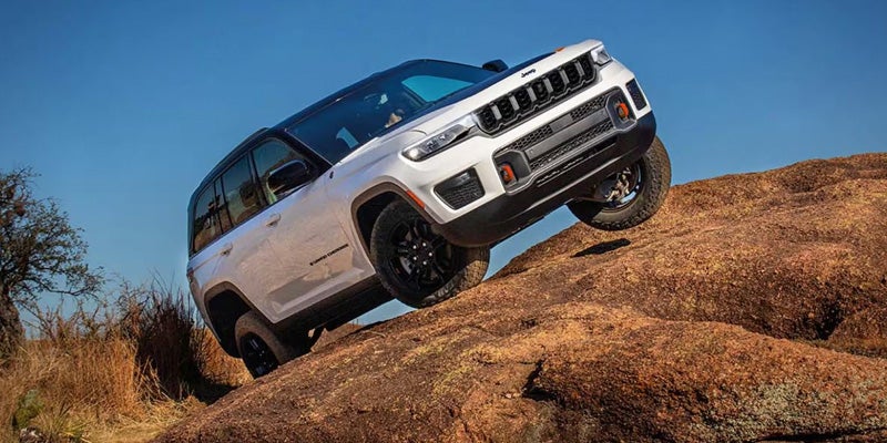 A white and black Jeep Grand Cherokee 4xe off-roading, with its front wheel lifted, on a rocky, rutted hillside.