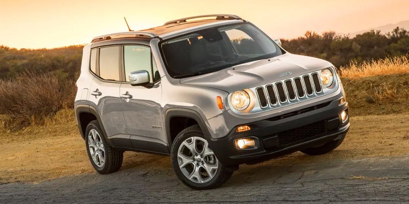 Silver Jeep Renegade SUV driving up a dirt path at sunset with headlights on.
