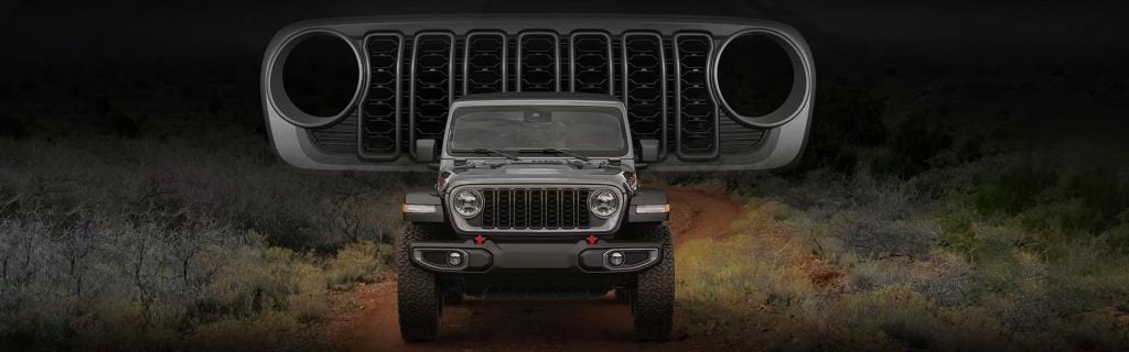 Front view of a gray Jeep Wrangler on a dirt track, featuring its grille and a large replica of the grille above it.