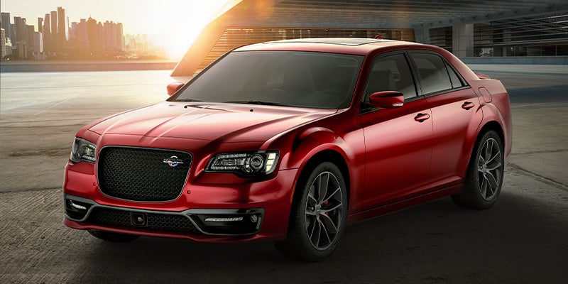 A glossy red Chrysler 300C parked on concrete, with a city skyline and sunset in the background.