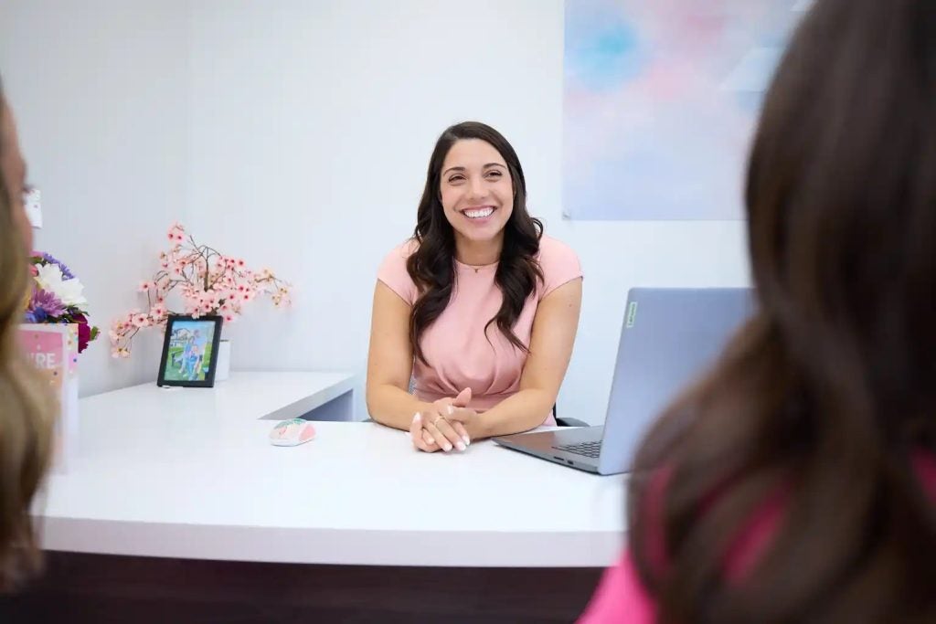 A smiling woman with long brown hair sits behind a white desk, conversing with someone whose back is to the camera.
