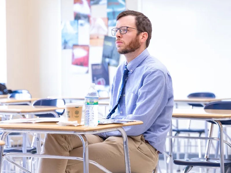 Teacher seated at a desk in a classroom, listening attentively