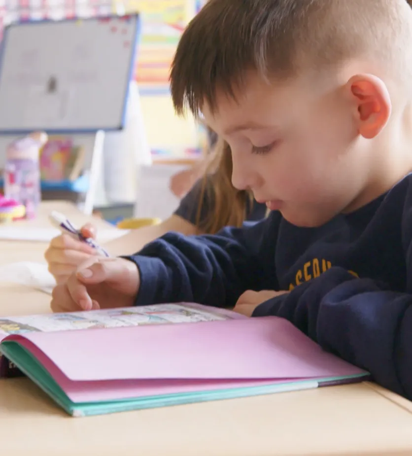 A young boy with short brown hair, wearing a navy blue sweatshirt, concentrates on drawing in an open book.