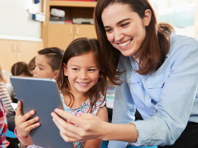 A smiling teacher and a young girl sit on a classroom floor together, looking at and interacting with a digital tablet.