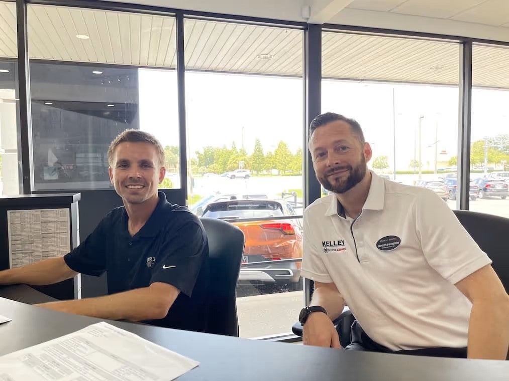 Two employees seated at a desk inside a car dealership office