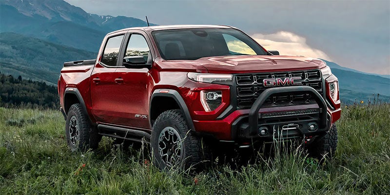 A red GMC pickup truck with a large black grille guard, light bar, and winch, parked in a grassy field with mountains.