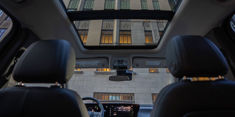 View from inside a car looking upward through a panoramic sunroof, showing a building exterior, with two front seats and a dashboard visible inside the vehicle.