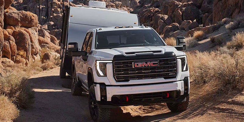 White GMC pickup truck on a dirt road in a rocky desert landscape, with large boulders and dry shrubs surrounding the vehicle and a trailer attached behind it.