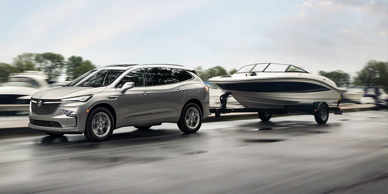 Silver Buick SUV towing a boat on a trailer along a wet road near a lakeside setting
