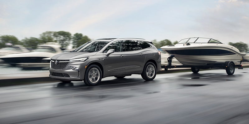 Silver SUV towing a boat on a trailer along a wet road, with blurred background vehicles and trees indicating motion under overcast daylight.