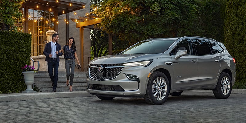 Silver SUV parked on a paved driveway outside a house with steps and string lights, with two people walking nearby and surrounded by trees and greenery at dusk.