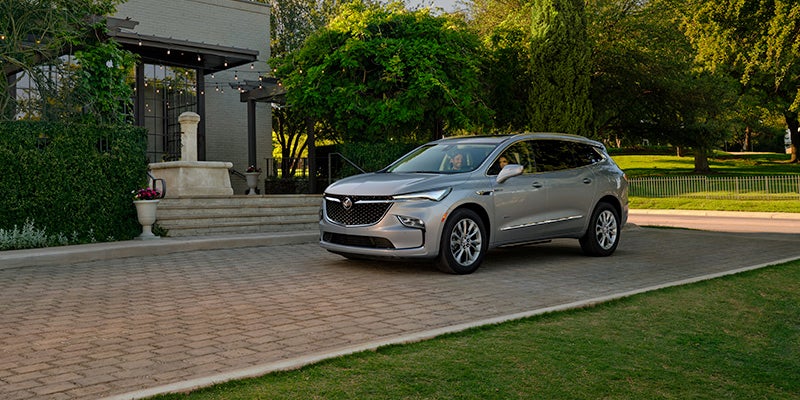 Silver minivan driving along a paved driveway past a house with a patio and steps, surrounded by lush greenery and trees in bright daylight