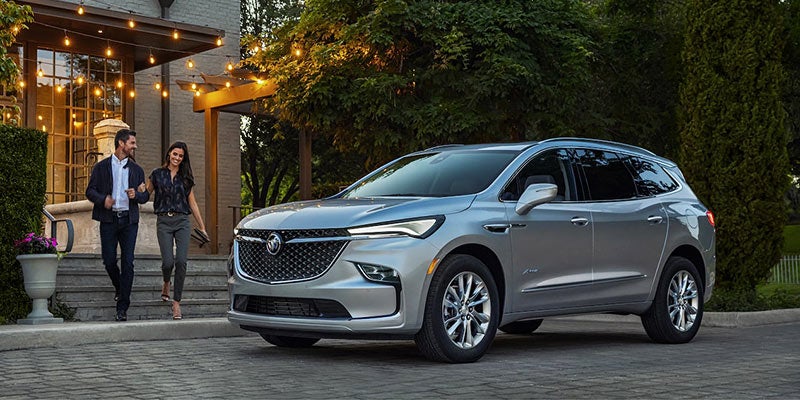 Silver Buick SUV parked in a driveway outside a house with string lights, with two people walking nearby.