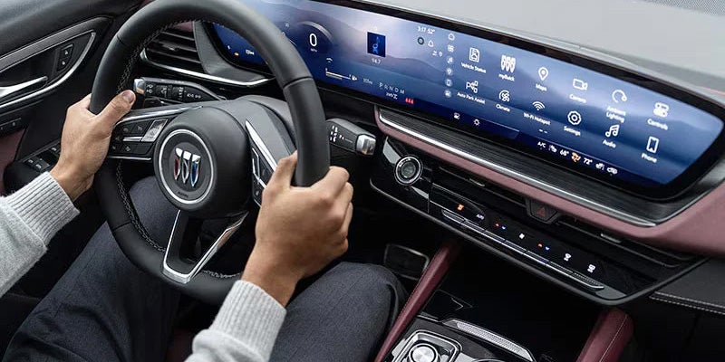 Hands on a steering wheel in a modern car with a wide digital display showing car settings and apps.