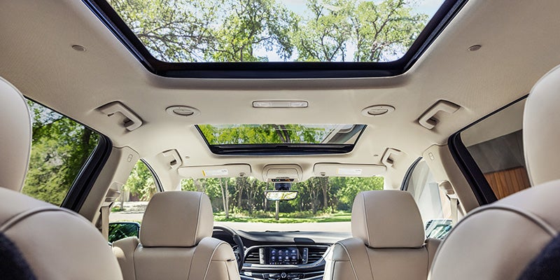Modern car interior with beige leather seats and a large panoramic sunroof, showing green trees and daylight outside