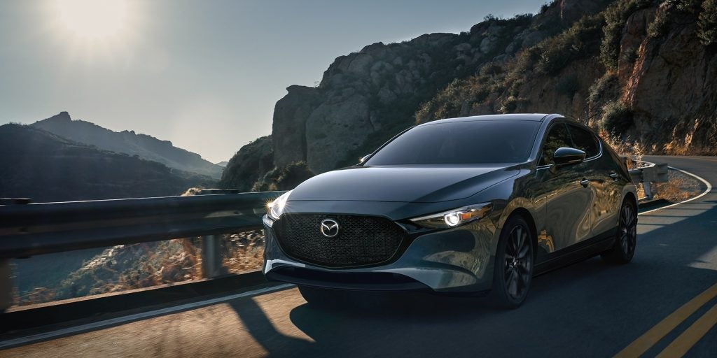 Gray Mazda3 sedan driving along a winding mountain road, showcasing its sleek exterior design, signature front grille