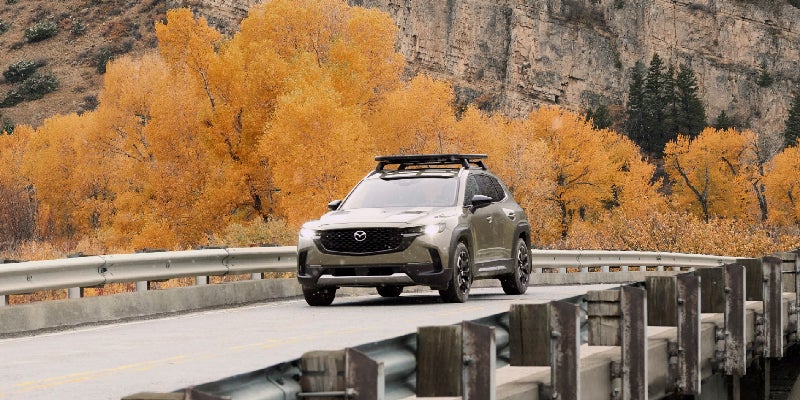 Gray Mazda CX-50 crossover SUV driving across a bridge surrounded by golden autumn foliage and rocky canyon walls