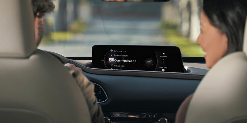 From behind, two people in a car look at a dashboard screen displaying "Communication" in a menu.