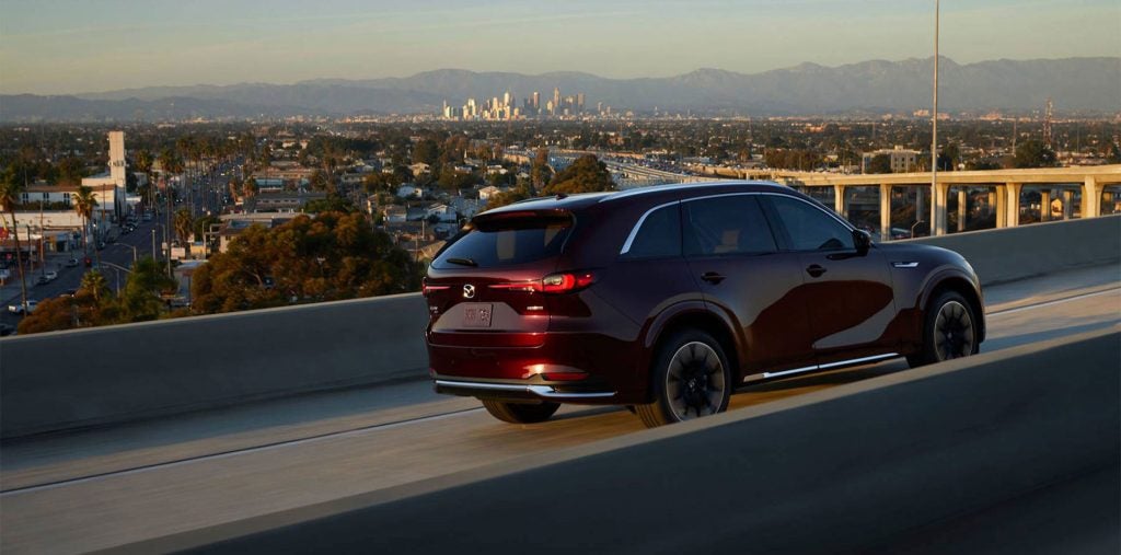 Rear three-quarter view of a dark red SUV driving on an elevated highway at sunset, with an urban skyline and distant mountains visible in the background.
