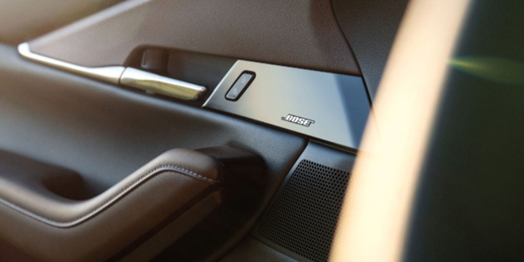 Close-up of a Mazda vehicle door panel featuring a Bose premium audio speaker, metallic trim, door handle, and stitched armrest, highlighting the vehicle’s upscale interior craftsmanship and sound system.