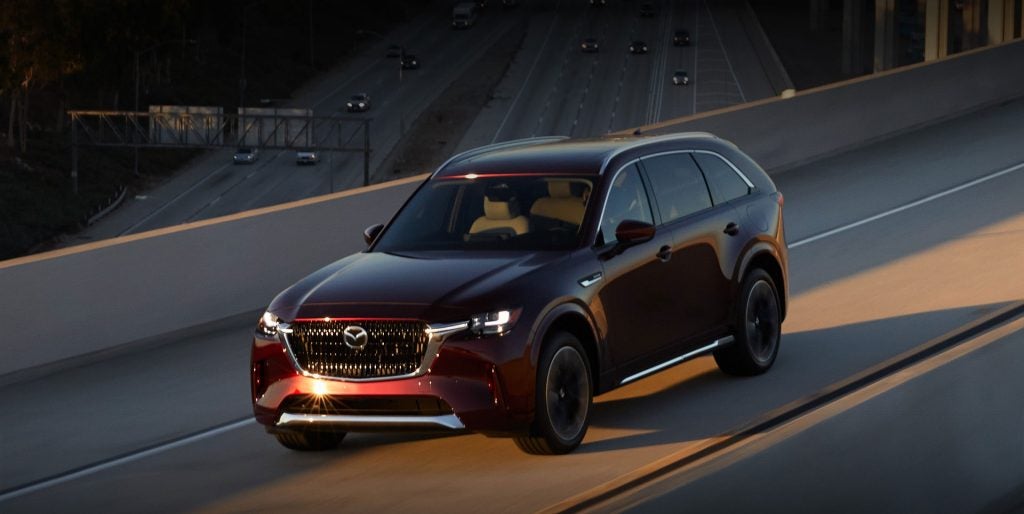 A dark red SUV travels along an elevated highway during dusk. The vehicle is viewed from the front-left angle