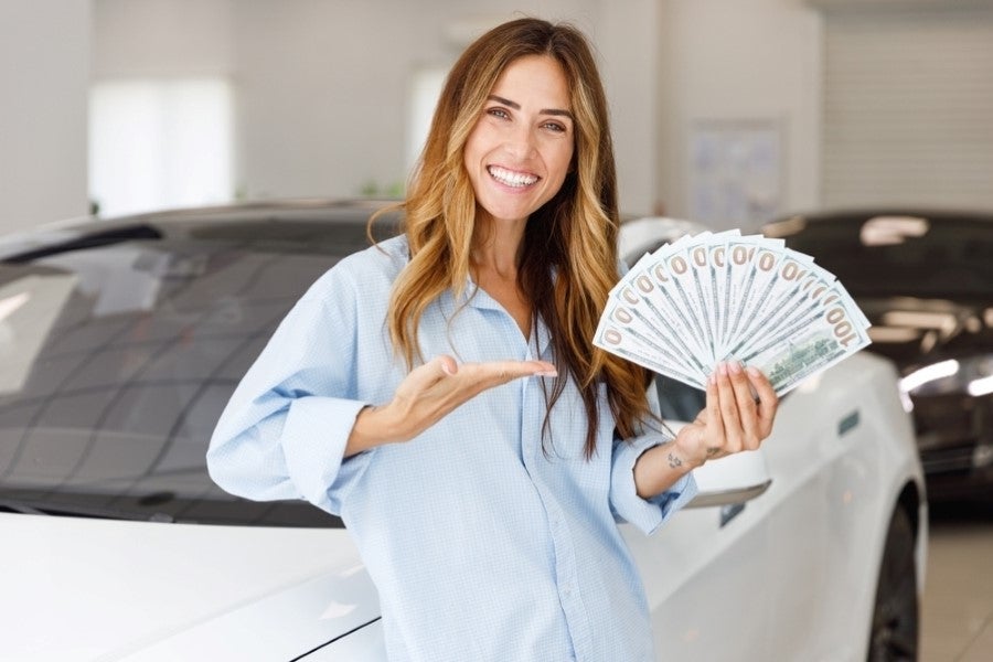 A smiling woman holding a fan of hundred-dollar bills and gesturing towards a white car beside her.