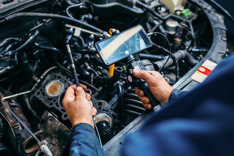 Mechanic inspecting a car engine with a borescope, viewing the internal components on a connected smartphone.