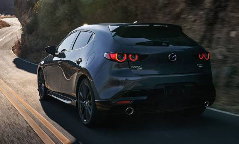 Rear three-quarter view of a dark Mazda hatchback driving along a winding mountain road, with rocky cliffs and sunlight in the background.