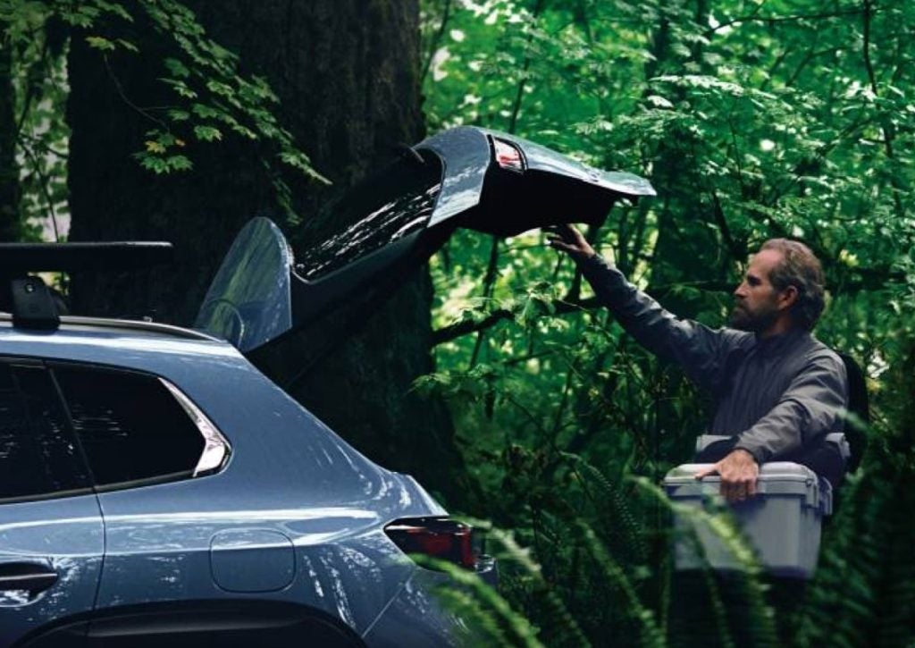 Person loading a portable cooler into the open rear cargo area of a Mazda SUV parked in a lush forest. The vehicle's liftgate is raised