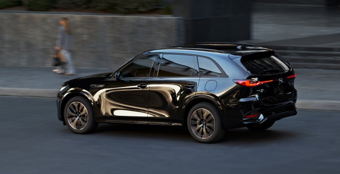 Black Mazda SUV driving on a city street, shown from a rear three-quarter angle with sleek body lines, tinted windows, and illuminated LED taillights.