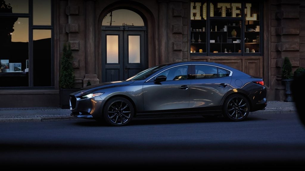 Side profile of a gray Mazda sedan parked along a city street at dusk, positioned in front of a building with illuminated hotel storefront windows.
