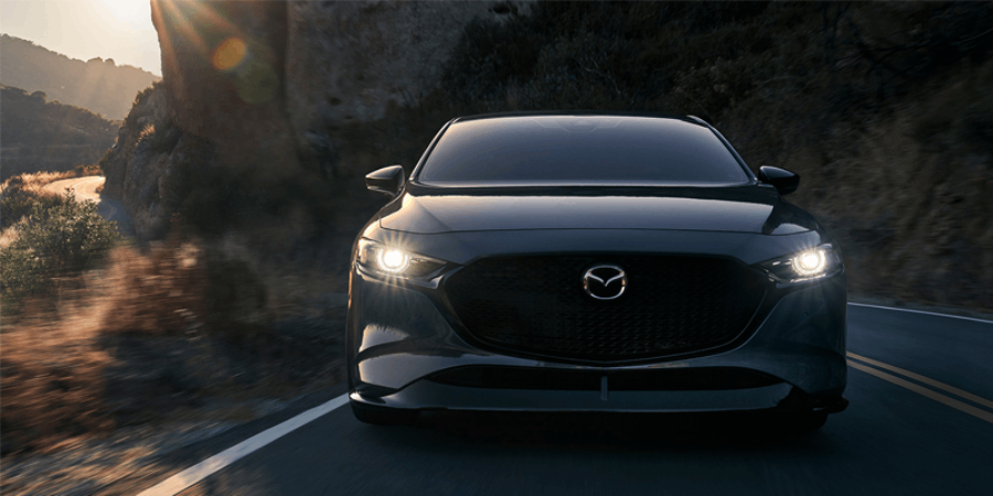 A dark gray car with its headlights on, driving on a winding road at dusk with hills in the background.