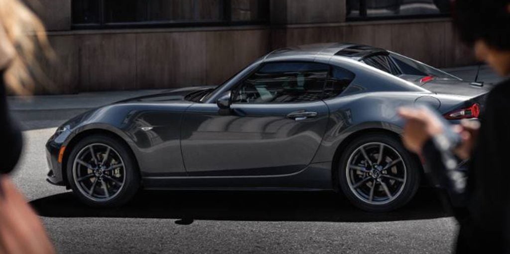 Side profile of a gray Mazda MX-5 Miata sports car parked on a city street, highlighting its sleek fastback-inspired silhouette