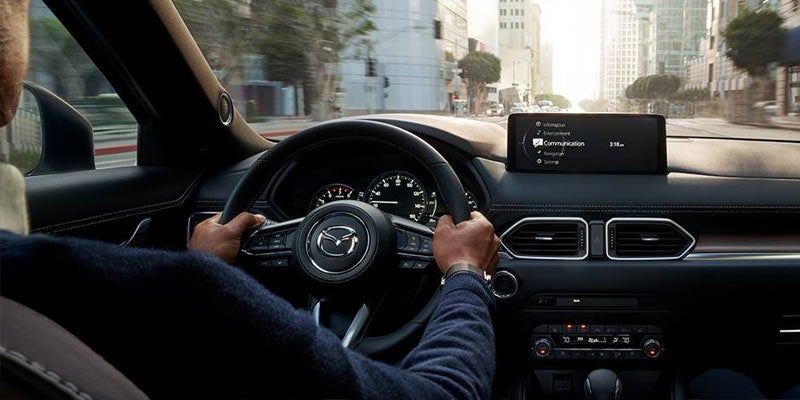 Driver's hands on a Mazda steering wheel, moving through a city with skyscrapers visible through the windshield.