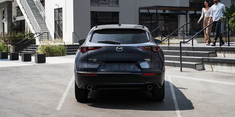 Rear view of a Mazda CX-5 parked in a marked parking space outside a modern residential building, with landscaped walkways, exterior staircases, and pedestrians visible in the background.
