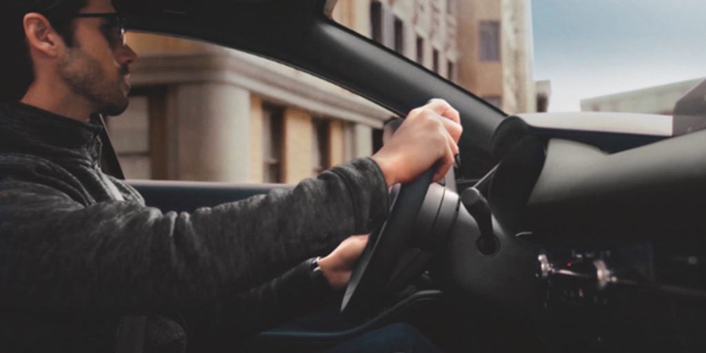 View from inside a Mazda vehicle showing a driver holding the steering wheel while traveling through an urban environment.