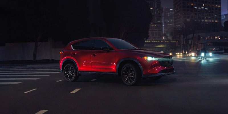 Red Mazda SUV driving through a city intersection at night, illuminated by streetlights and surrounding building lights.