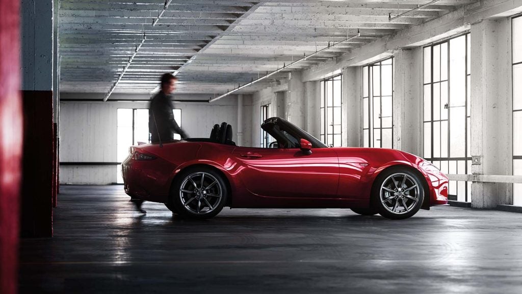 A man approaches a shiny red convertible Mazda Miata parked in a concrete garage with tall windows.