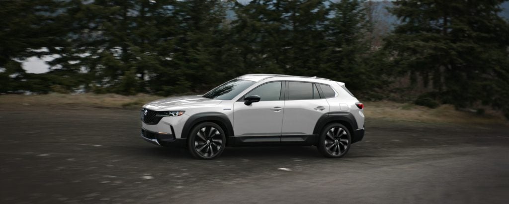 Side profile of a white Mazda SUV driving on a paved road through a wooded landscape. Motion blur in the background emphasizes movement