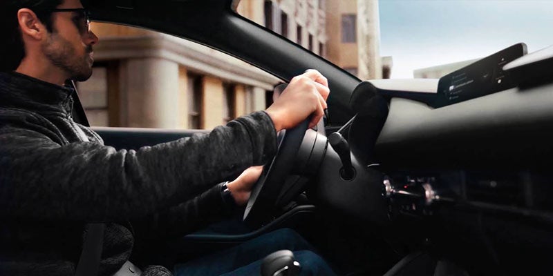 Driver seated behind the wheel of a Mazda vehicle, highlighting the cockpit-style interior, steering controls, and dashboard design while driving in an urban setting.