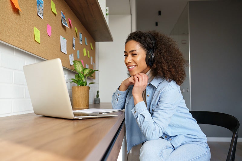 Smiling woman with headphones watches a laptop on a wooden desk with a corkboard and plant.