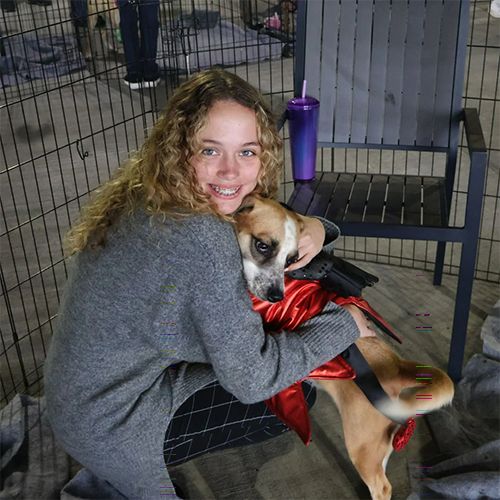 Smiling girl hugging a dog dressed in a red outfit while sitting indoors.